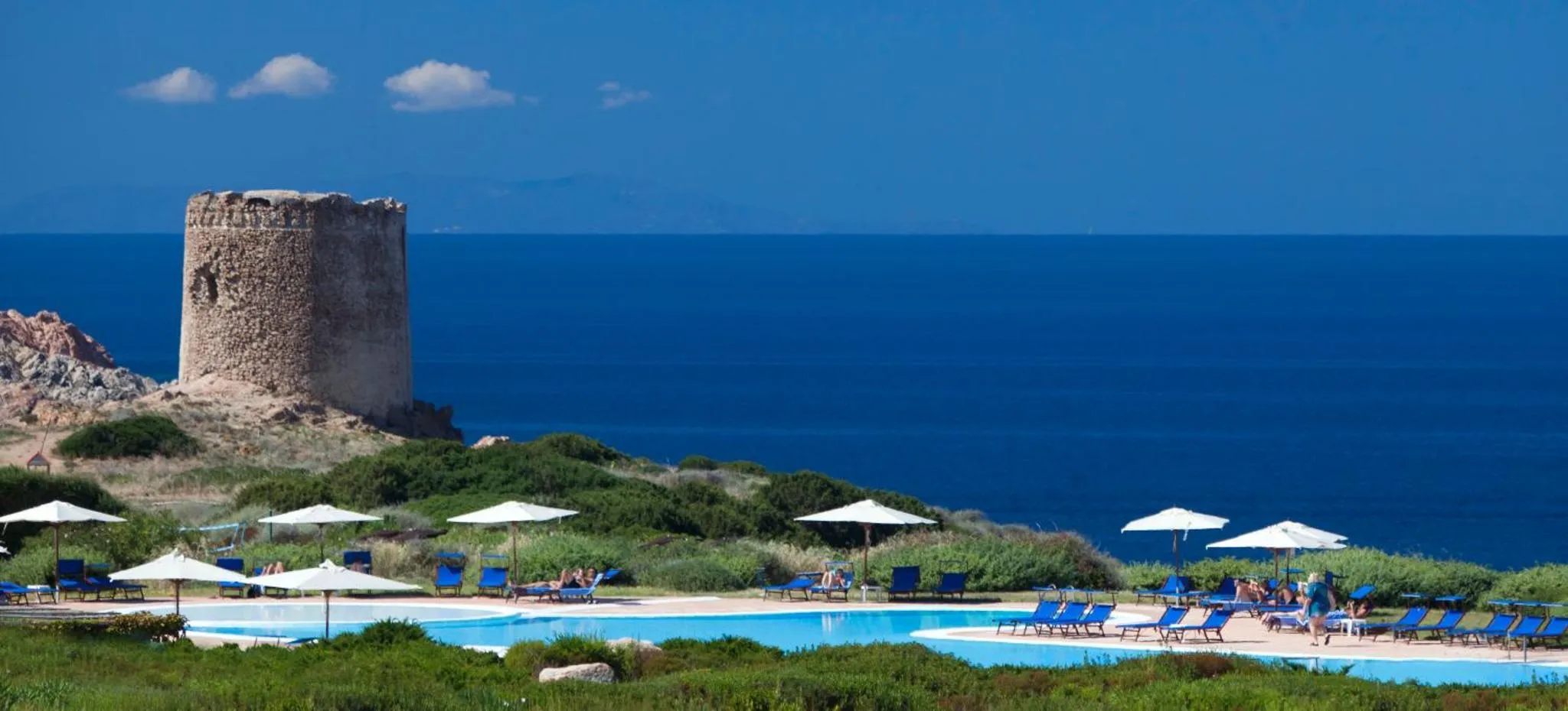 Une piscine bordée de transats et de parasols offre une vue sur la mer et une tour côtière.