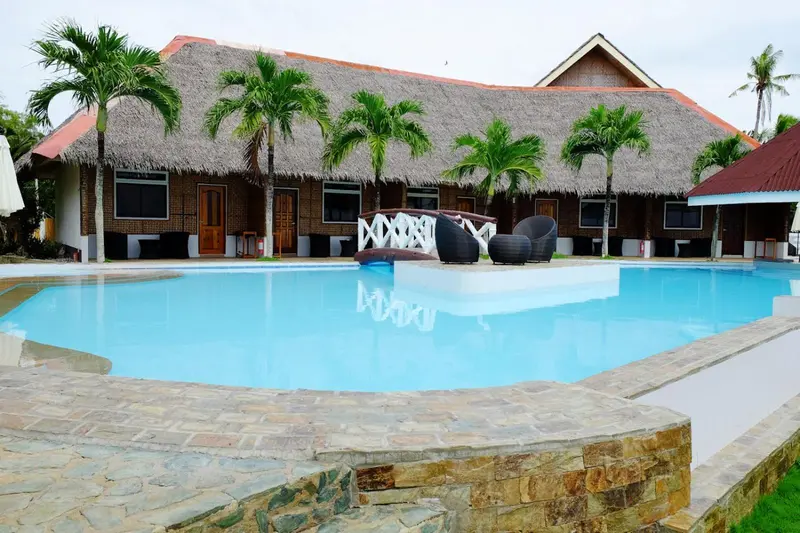 Une piscine turquoise bordée d'une terrasse en pierre, devant un bâtiment à toit de chaume.
