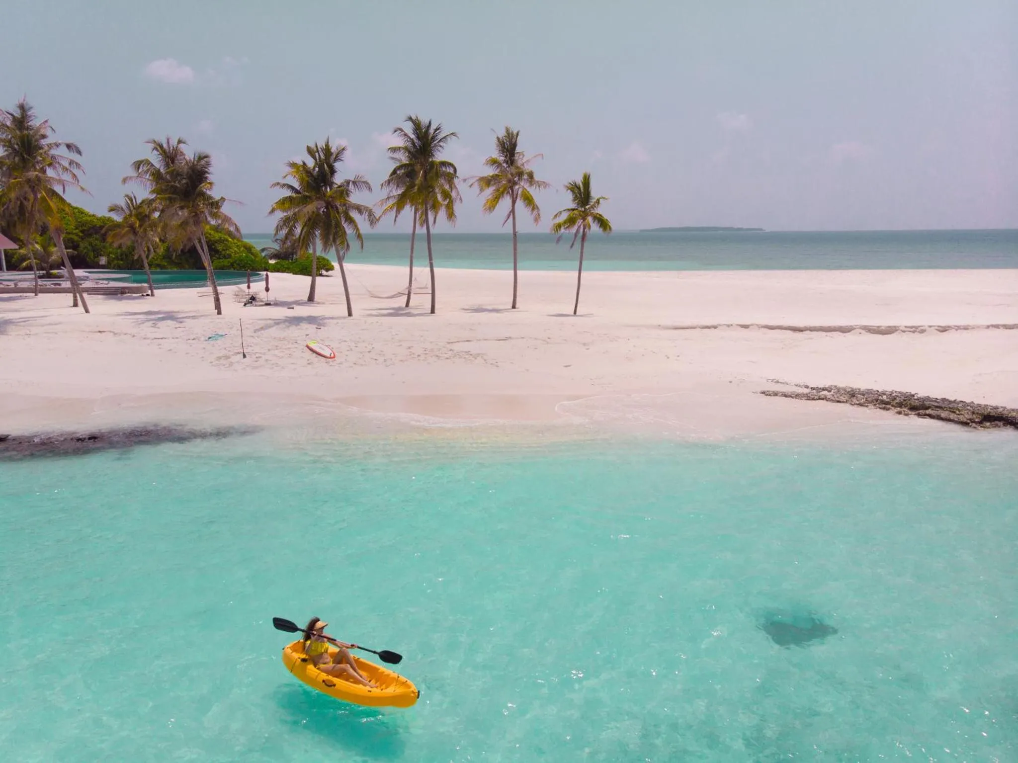 Une personne en kayak navigue sur une eau turquoise peu profonde près d'une plage de sable blanc bordée de palmiers.