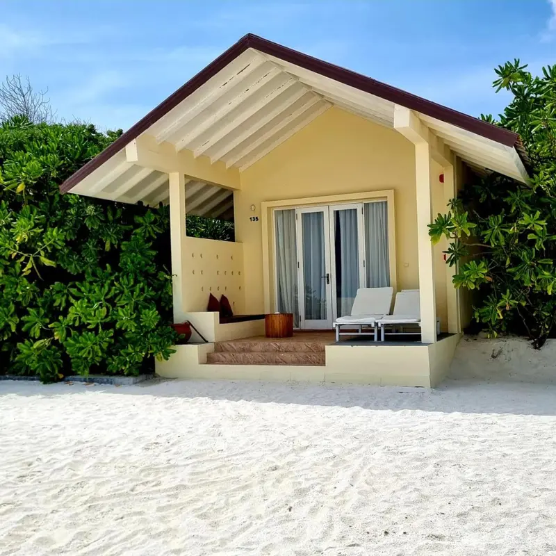 Une petite maison jaune avec une terrasse donnant sur une plage de sable blanc.
