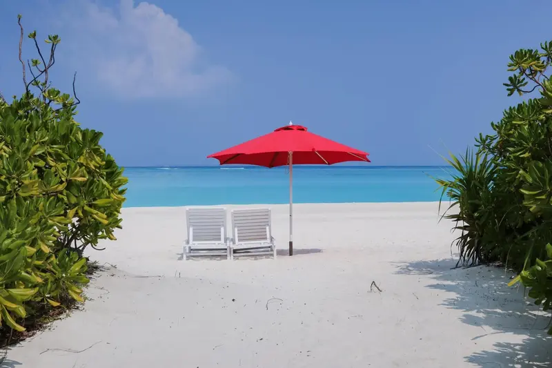 Deux chaises longues blanches sous un parasol rouge sur une plage de sable blanc.