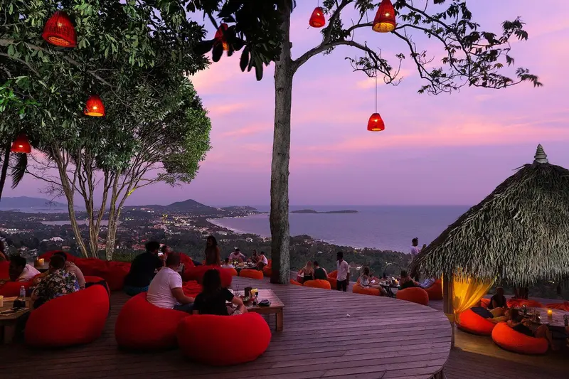 Terrasse avec mobilier rouge et vue sur un paysage côtier au coucher du soleil.