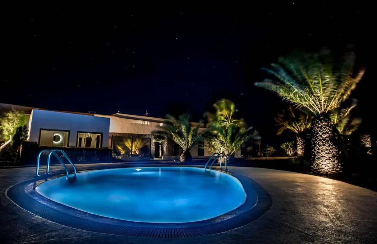 Une piscine illuminée au premier plan, avec un bâtiment et des palmiers en arrière-plan sous un ciel étoilé.