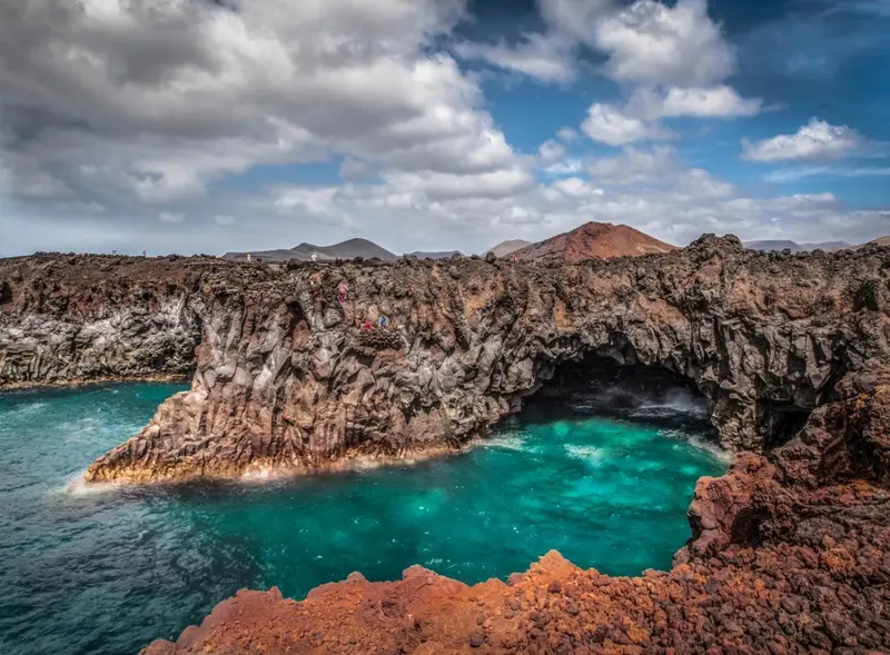Côte rocheuse volcanique surplombant une crique turquoise sous un ciel nuageux.