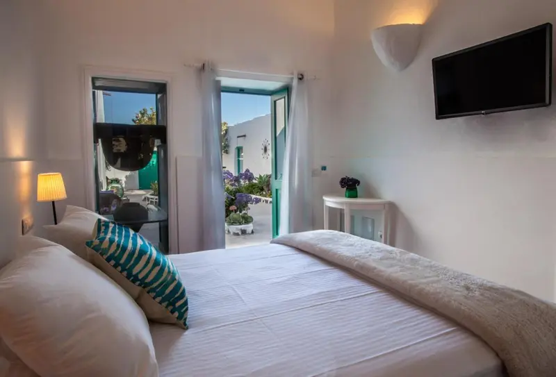 Une chambre lumineuse avec un lit recouvert d'une couverture blanche et un oreiller turquoise, donnant sur une cour verdoyante.