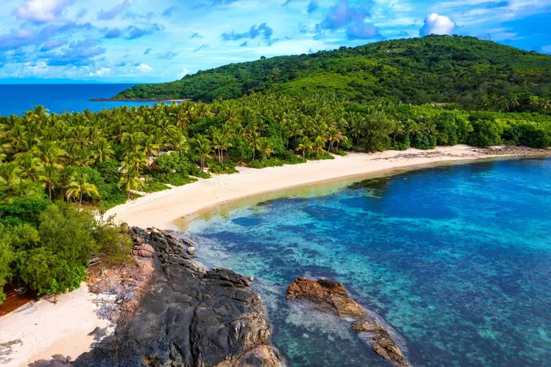 Vue aérienne d'une plage de sable blanc bordée de végétation luxuriante et d'eaux turquoise.