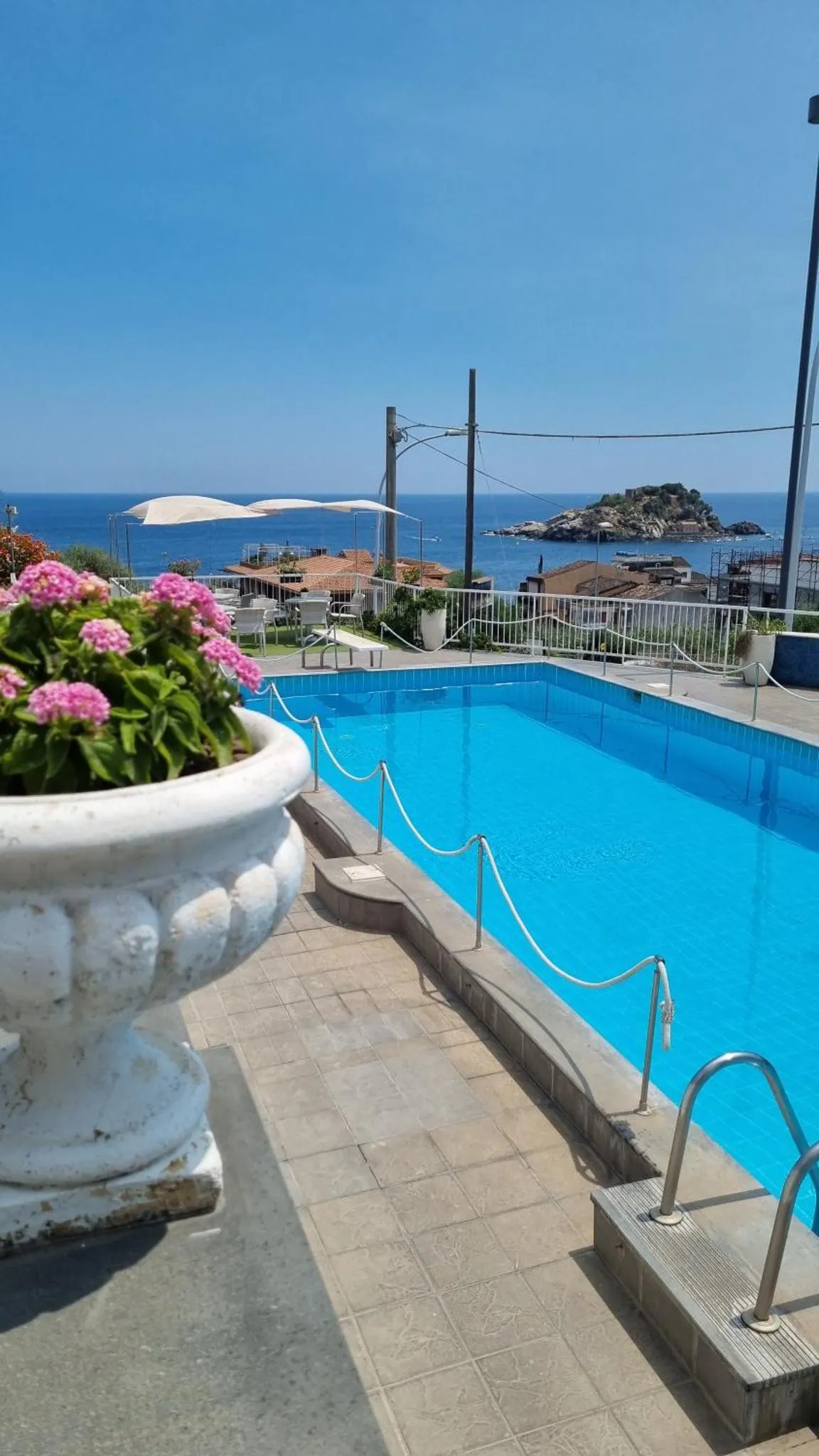 Une piscine turquoise bordée d'une terrasse ensoleillée offre une vue sur la mer et un îlot rocheux.