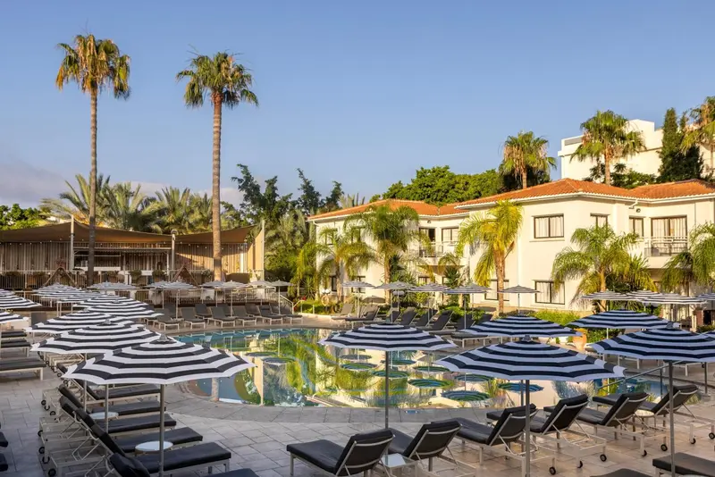 Transats et parasols rayés bordent une piscine rectangulaire dans un jardin tropical.