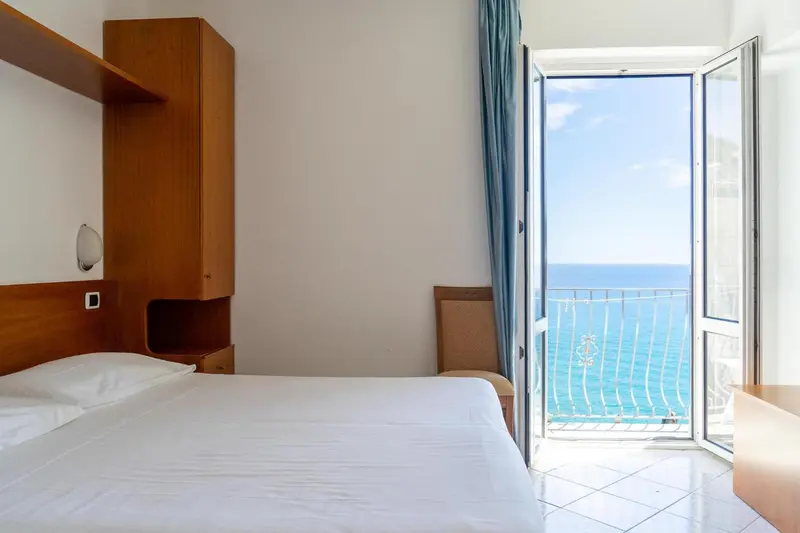 Chambre lumineuse avec un lit simple et une vue sur la mer depuis un balcon.