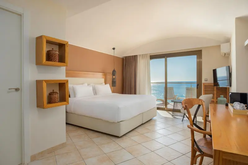 Une chambre lumineuse avec un grand lit face à une baie vitrée donnant sur la mer.