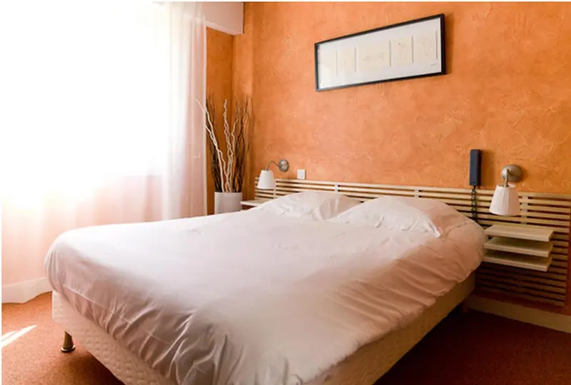 Une chambre lumineuse avec un lit recouvert de draps blancs et un mur orange texturé.