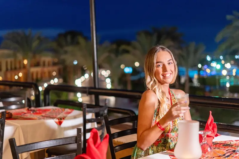 Une femme souriante est assise à une table de restaurant en extérieur, avec une boisson à la main.