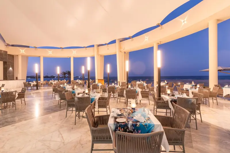 Une salle de restaurant lumineuse avec des tables dressées face à la mer au crépuscule.