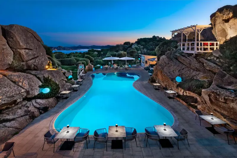 Piscine illuminée avec tables et chaises, au milieu d'un paysage rocheux et végétal.