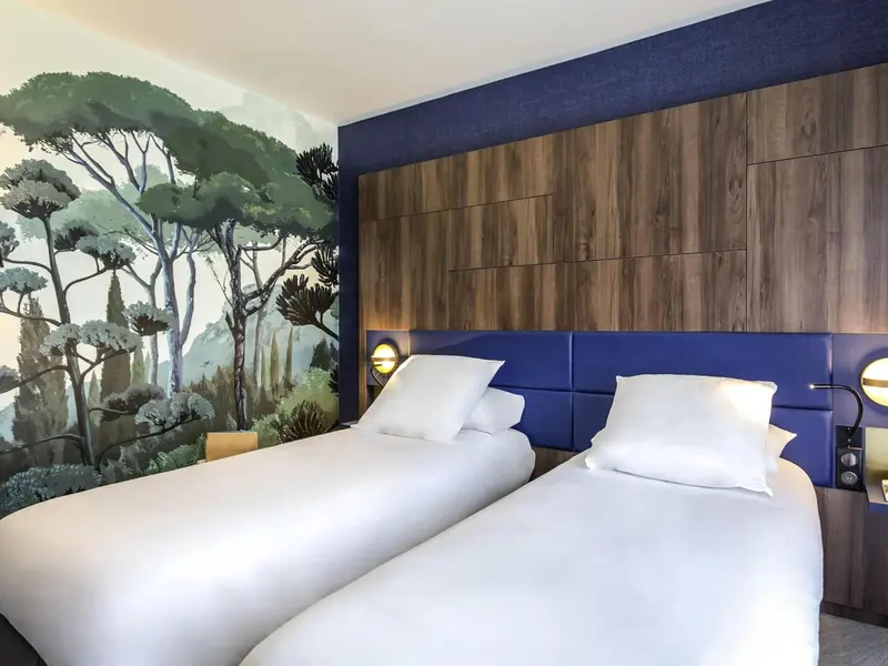Deux lits simples avec oreillers blancs dans une chambre décorée d'un mur illustré d'une forêt.