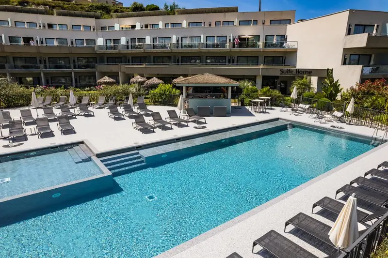 Piscine rectangulaire entourée de transats et de végétation luxuriante devant un bâtiment blanc.