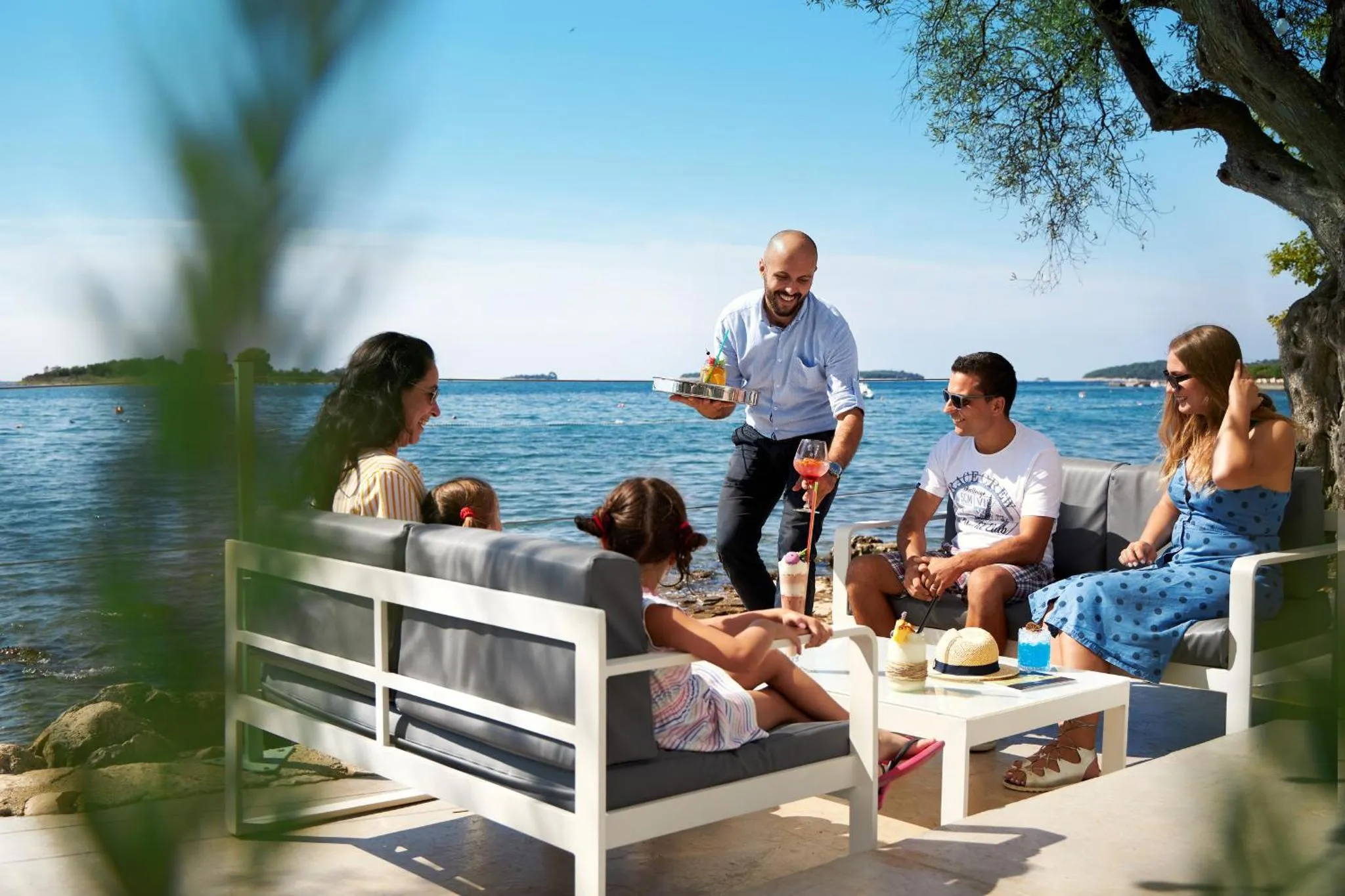 Des clients se détendent sur un canapé en extérieur face à la mer, tandis qu'un serveur leur apporte des boissons.