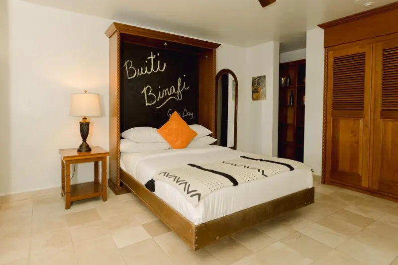Une chambre lumineuse avec un grand lit recouvert d'une courtepointe blanche et d'un oreiller orange.