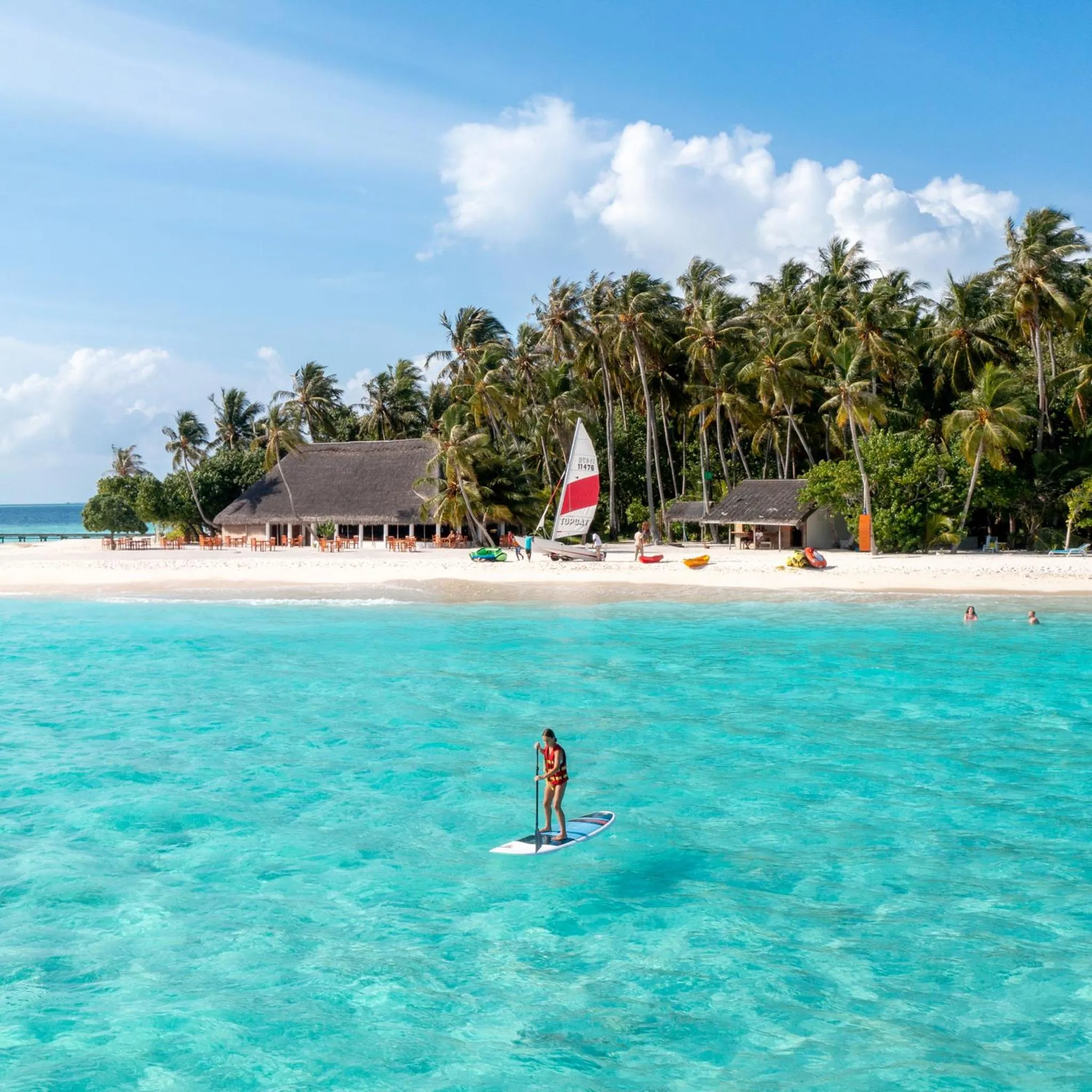 Une personne pratique le stand-up paddle dans une eau turquoise face à une plage de sable blanc bordée de palmiers.
