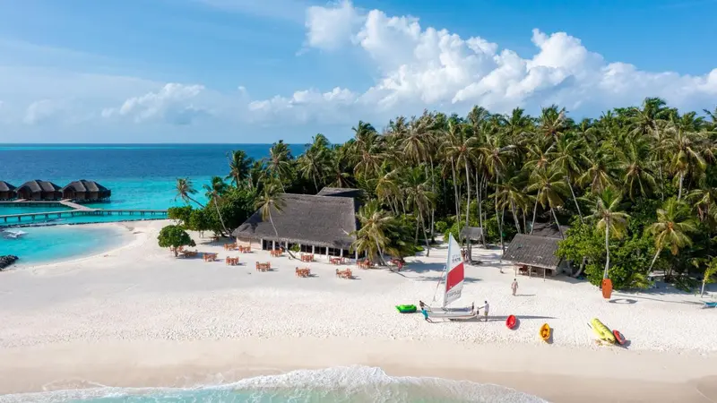 Vue aérienne d'une plage de sable blanc bordée de végétation luxuriante et de bungalows sur pilotis.