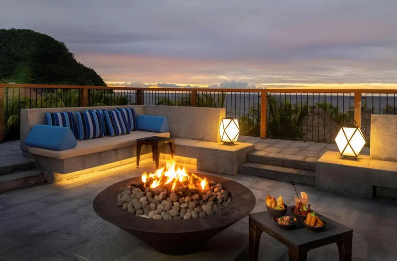 Un espace de détente extérieur avec un foyer central et une vue sur la mer au coucher du soleil.