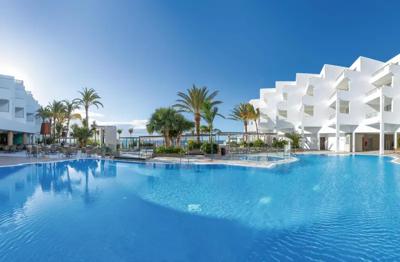 Vue d'une piscine turquoise devant un bâtiment blanc moderne et des palmiers.