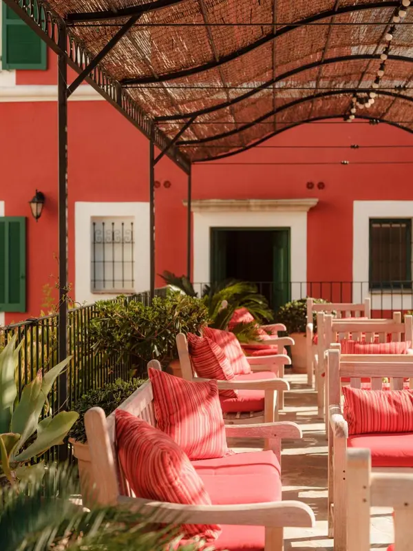 Terrasse ombragée avec des bancs en bois et des coussins rouges devant un bâtiment coloré.