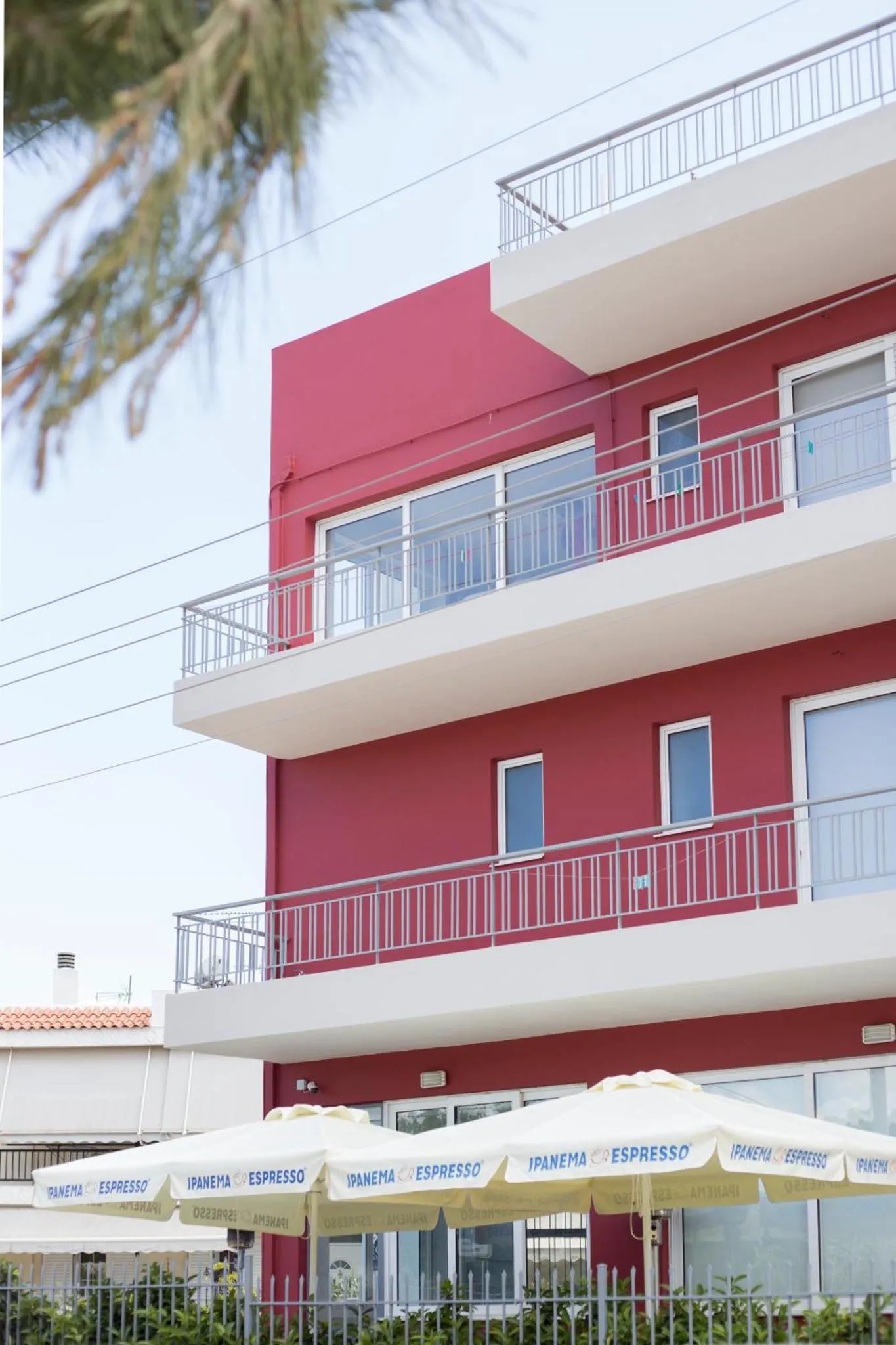 Façade d'un bâtiment rouge à balcons blancs surplombant une terrasse ombragée.