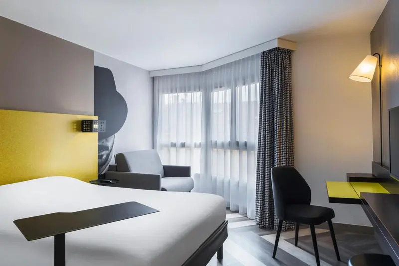 Une chambre d'hôtel moderne avec un lit, un bureau et un fauteuil près d'une fenêtre.