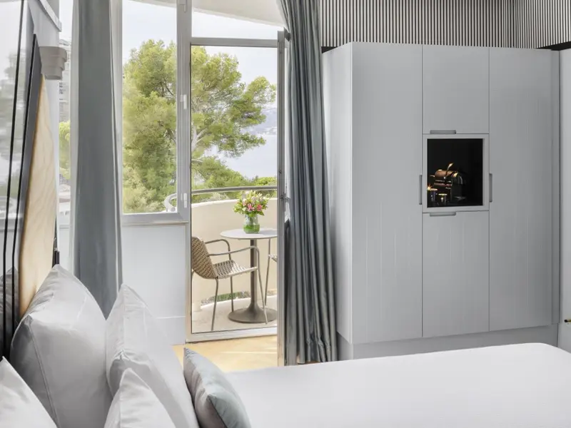 Une chambre lumineuse s'ouvre sur un balcon avec vue sur la végétation et la mer.