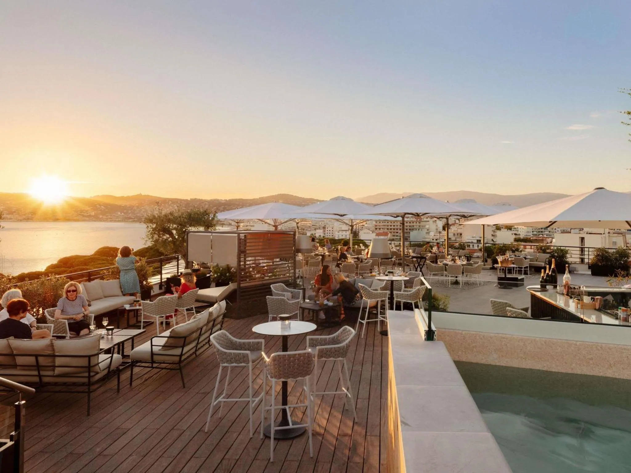 Terrasse aménagée avec tables et chaises, offrant une vue sur la mer et le coucher de soleil.