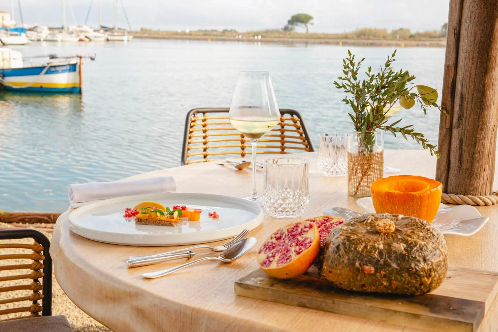 Une table est dressée avec un plat de poisson, des fruits et des verres sur une terrasse en bord de mer.