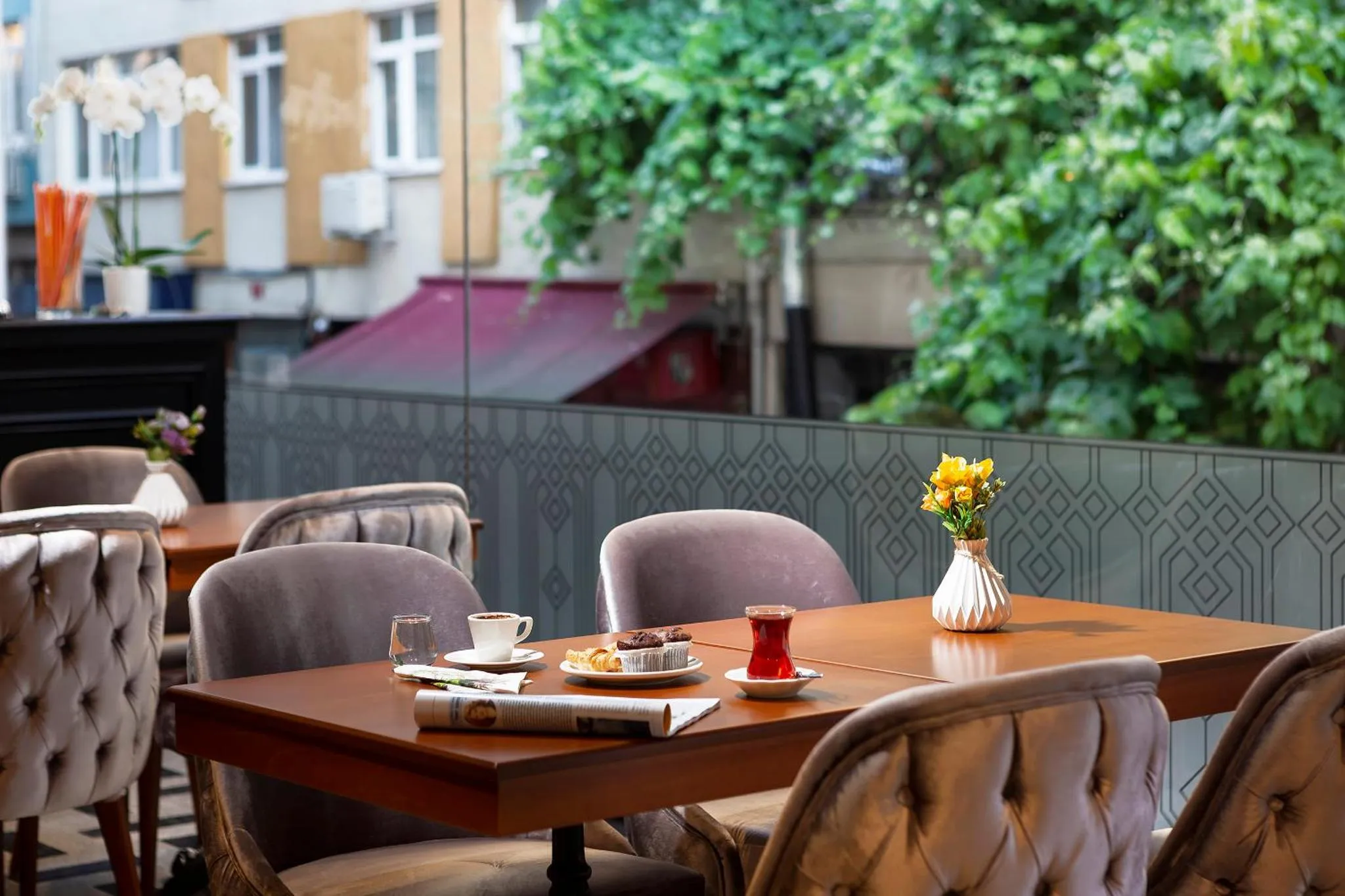 Une table est dressée avec une tasse de thé, des pâtisseries et un journal sur une terrasse ombragée.