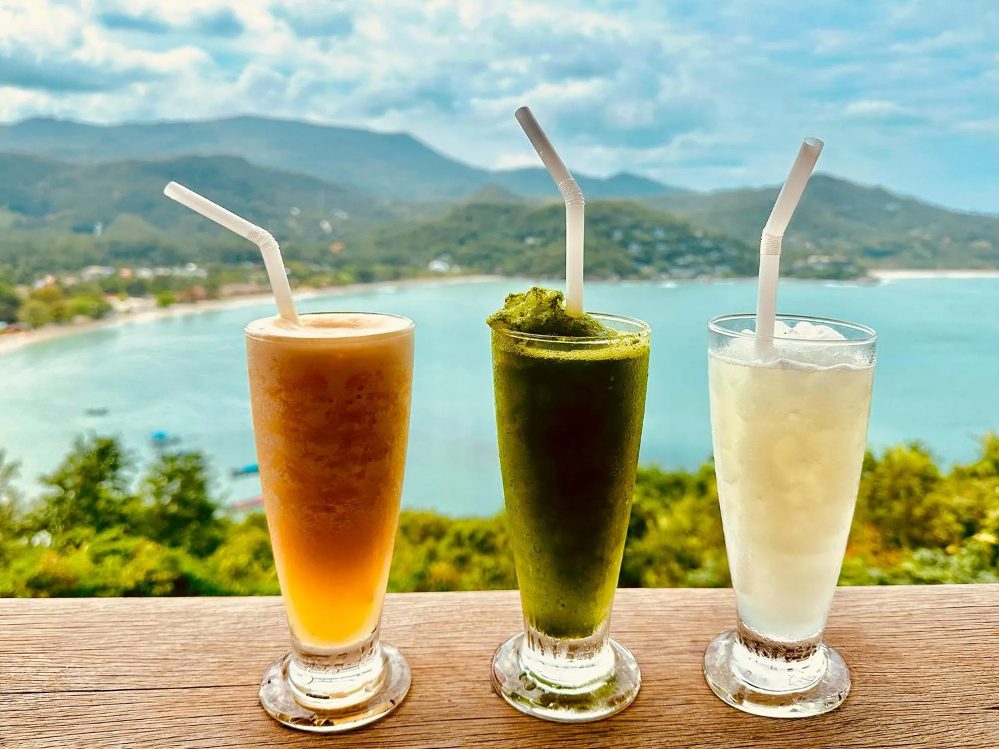 Trois verres de boissons colorées sur une table en bois avec vue sur la mer et les montagnes.