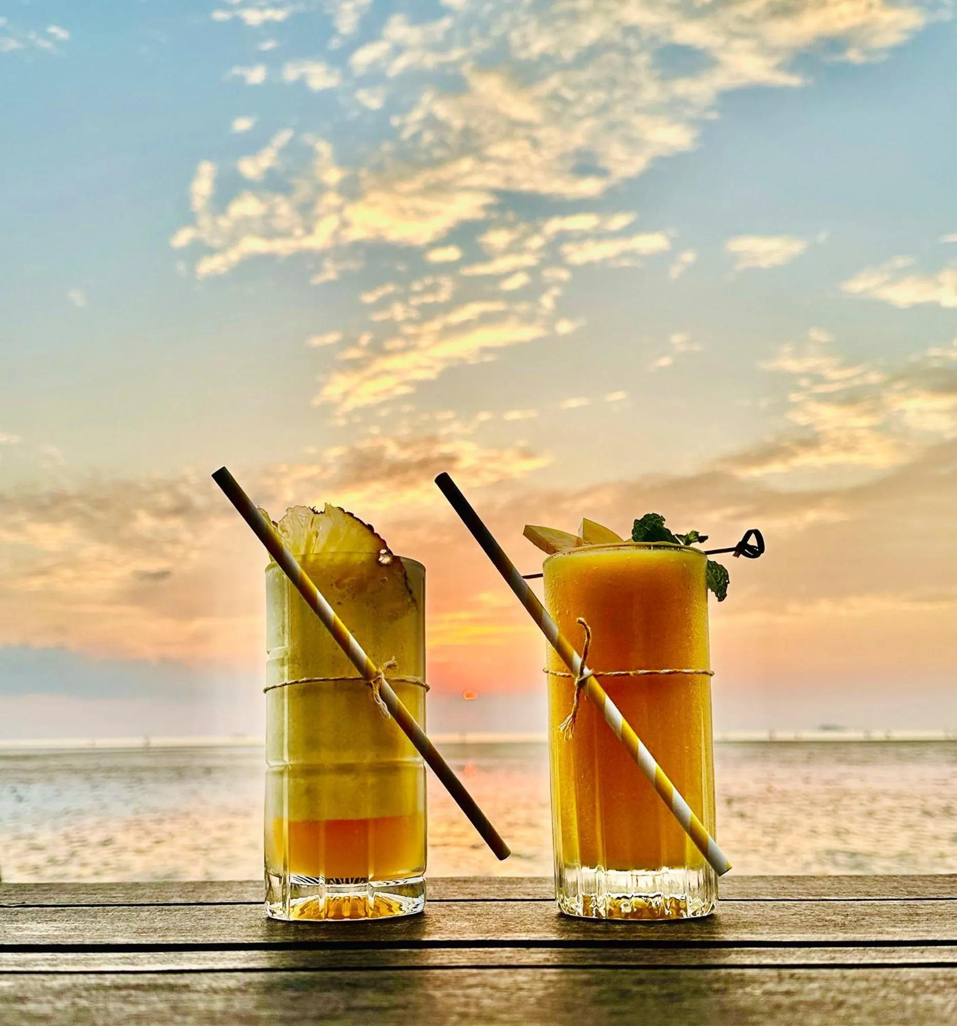Deux cocktails exotiques sont servis sur une terrasse en bois face à la mer au coucher du soleil.