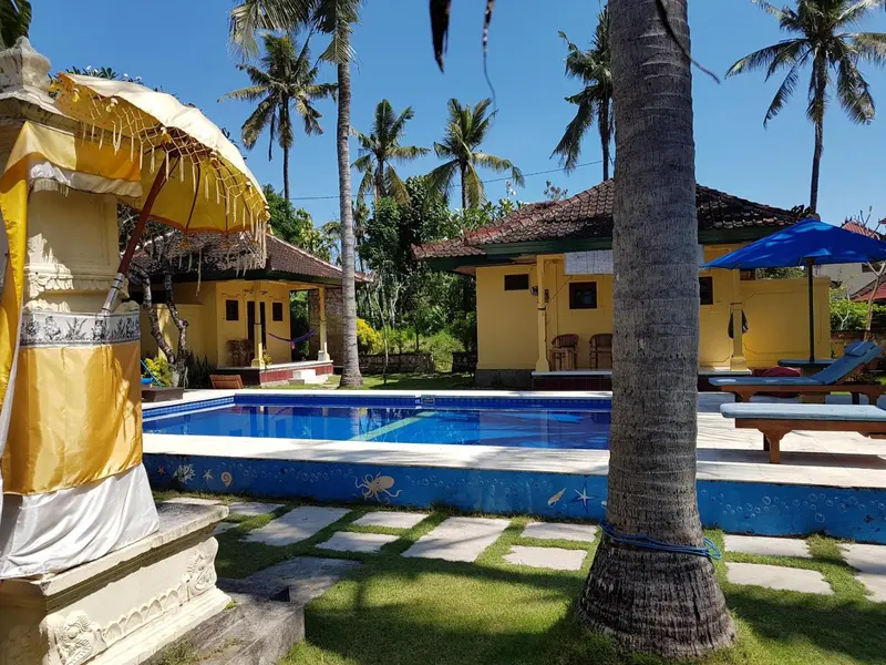 Une piscine rectangulaire au premier plan, entourée d'une pelouse verdoyante et de bâtiments jaunes.