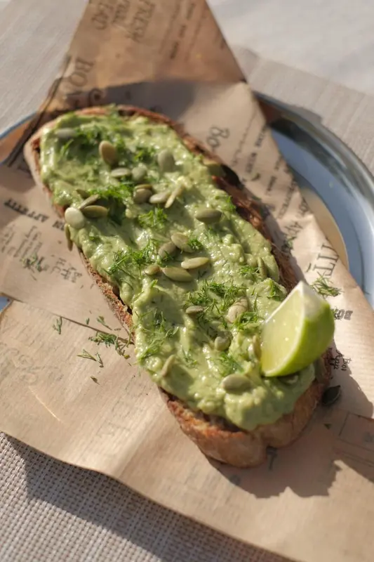 Une tartine à l'avocat, décorée de graines et d'une tranche de citron vert, est présentée sur du papier.