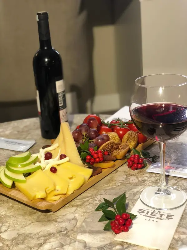 Une table présente une bouteille de vin rouge, un verre rempli, et une assiette de fromages et de fruits.