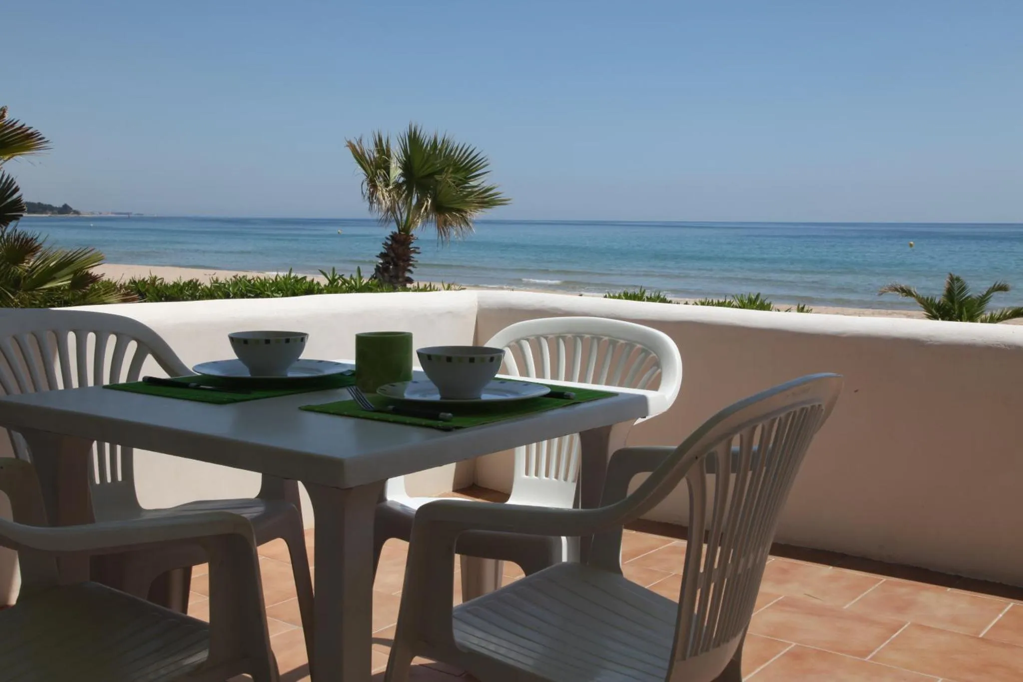 Une table et des chaises en plastique blanc sont dressées sur une terrasse carrelée face à la mer.