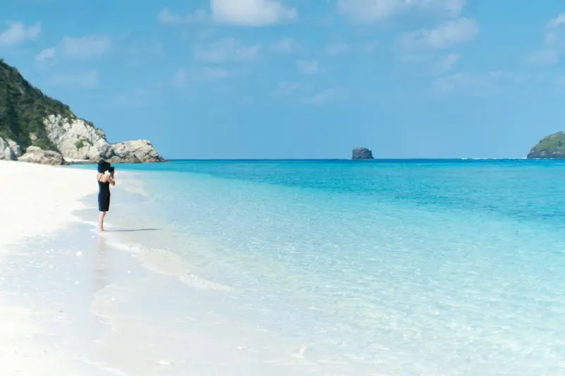 Une personne se tient sur une plage de sable blanc face à une mer turquoise.