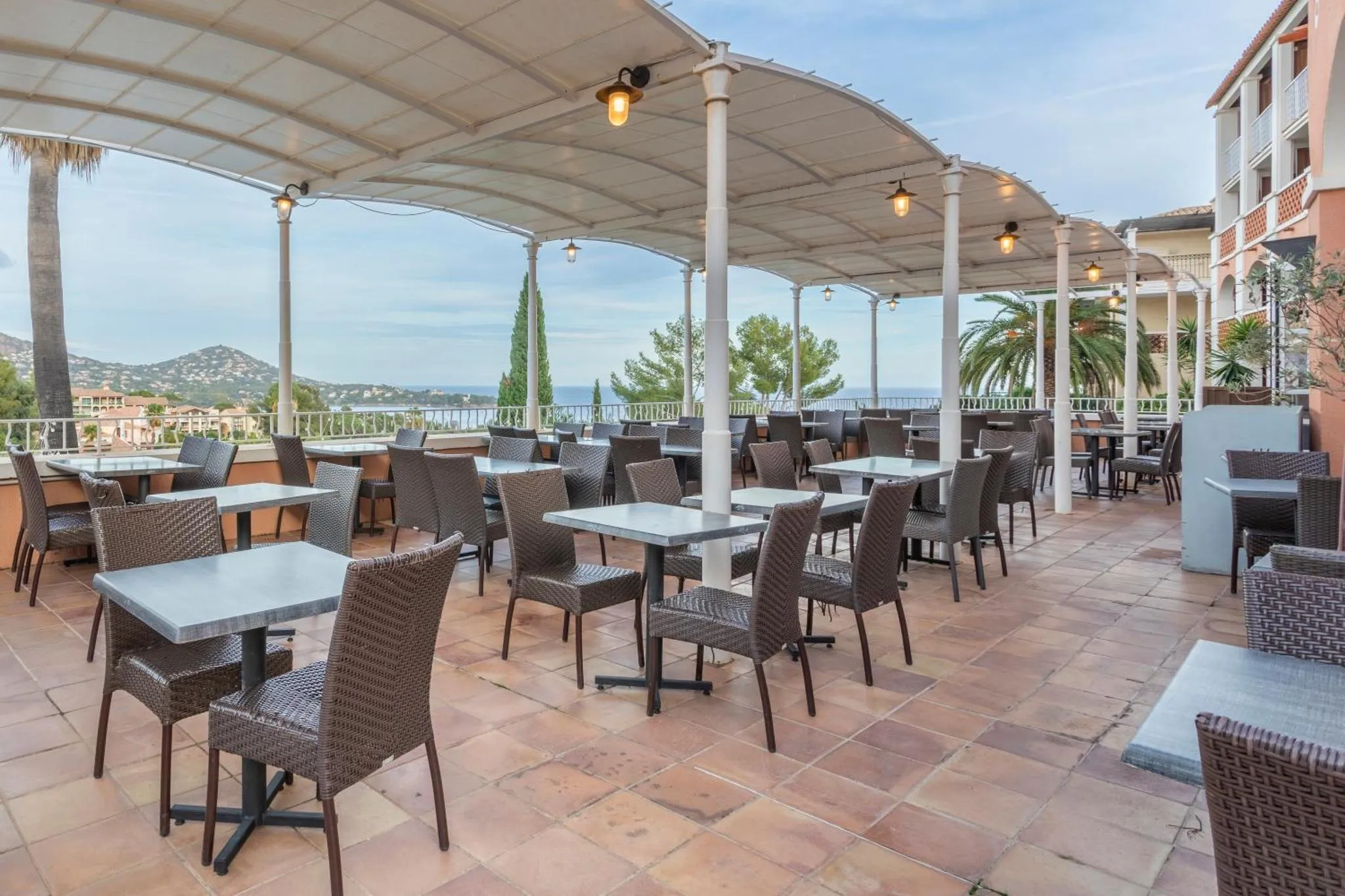 Terrasse ombragée avec tables et chaises donnant sur la mer et les collines.