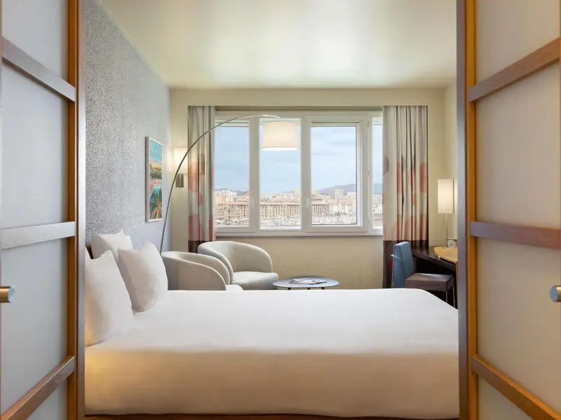Une chambre lumineuse avec un lit et un coin salon donnant sur une vue urbaine.