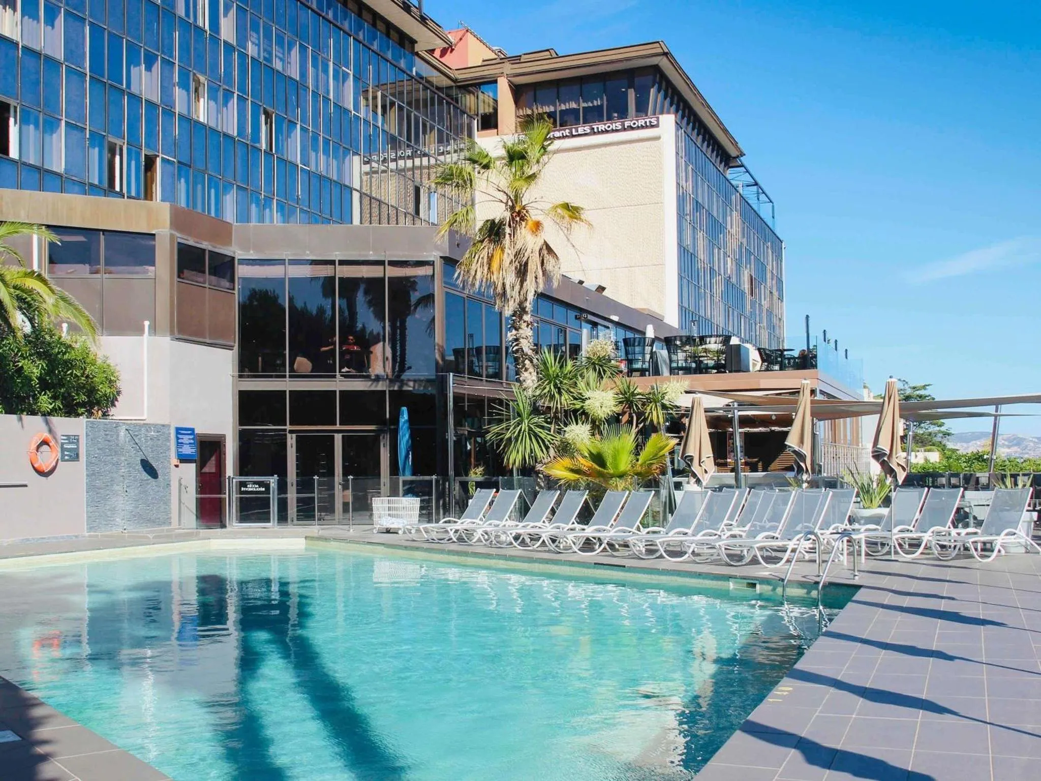 Piscine d'hôtel avec transats et palmiers sous un ciel bleu.