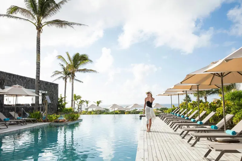 Une femme se tient près d'une piscine bordée de transats sous des parasols.