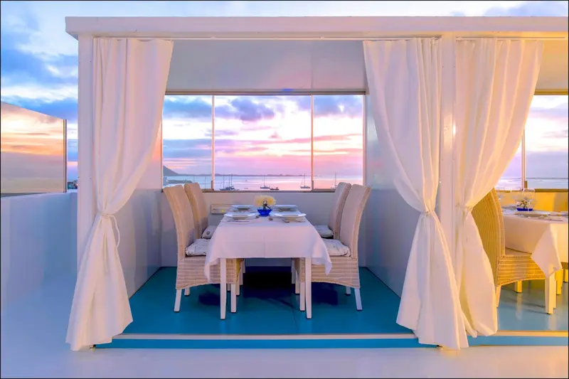Une table est dressée dans un espace extérieur couvert avec vue sur la mer au coucher du soleil.