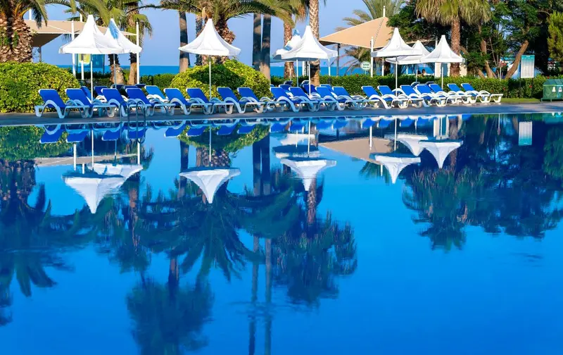 Transats bleus alignés au bord d'une piscine turquoise reflétant les palmiers et les parasols.
