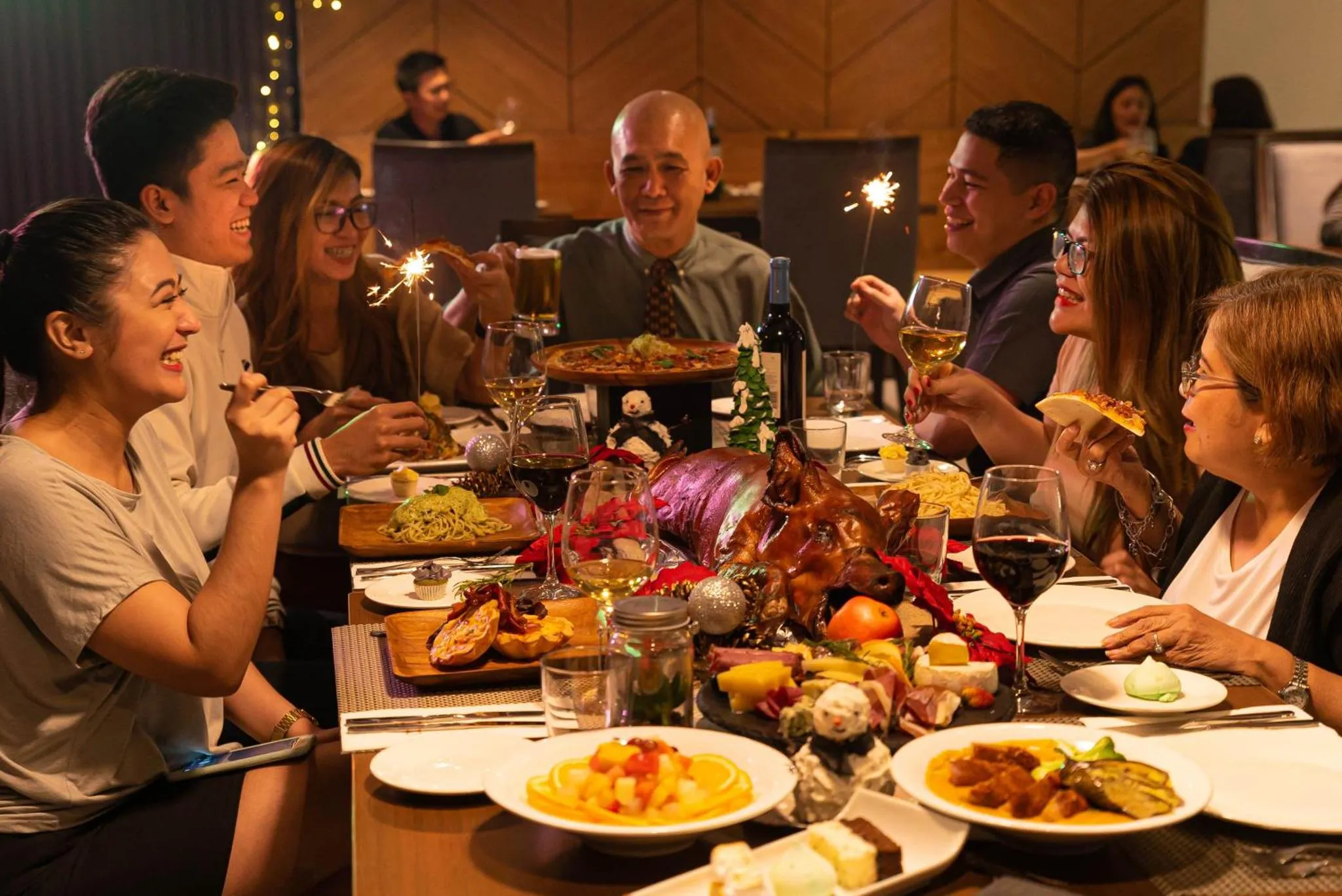 Une table festive est garnie de plats variés et de boissons, autour de laquelle sont assis des convives souriants.
