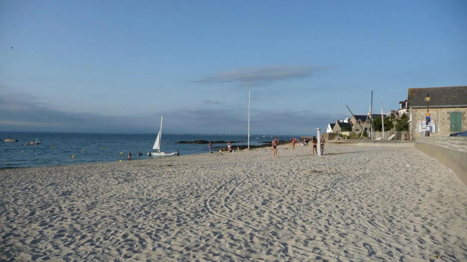 Plage de sable fin avec des personnes se promenant et des voiliers en mer.