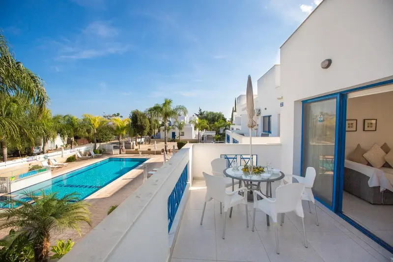Terrasse avec table et chaises devant une piscine et un bâtiment blanc.