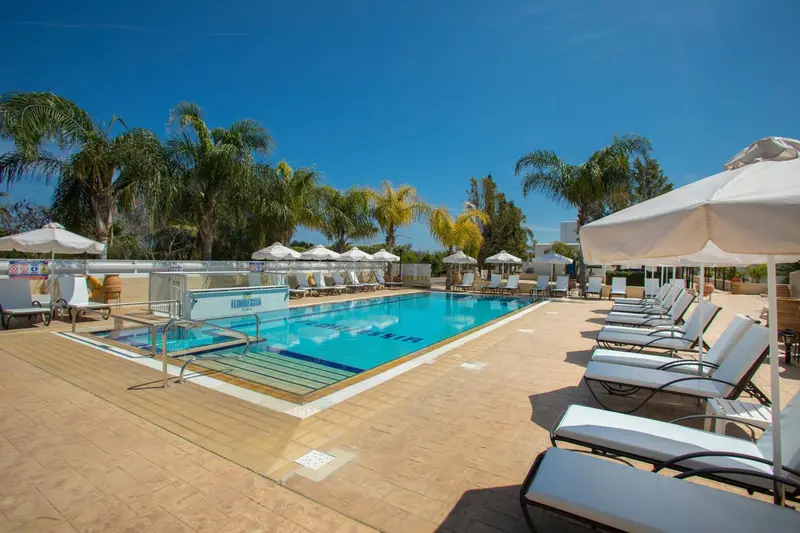 Transats blancs alignés au bord d'une piscine rectangulaire sous un ciel bleu.